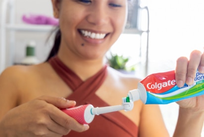 Una mujer sonríe mientras pone pasta de dientes Colgate en un cepillo de dientes en un cuarto de baño.